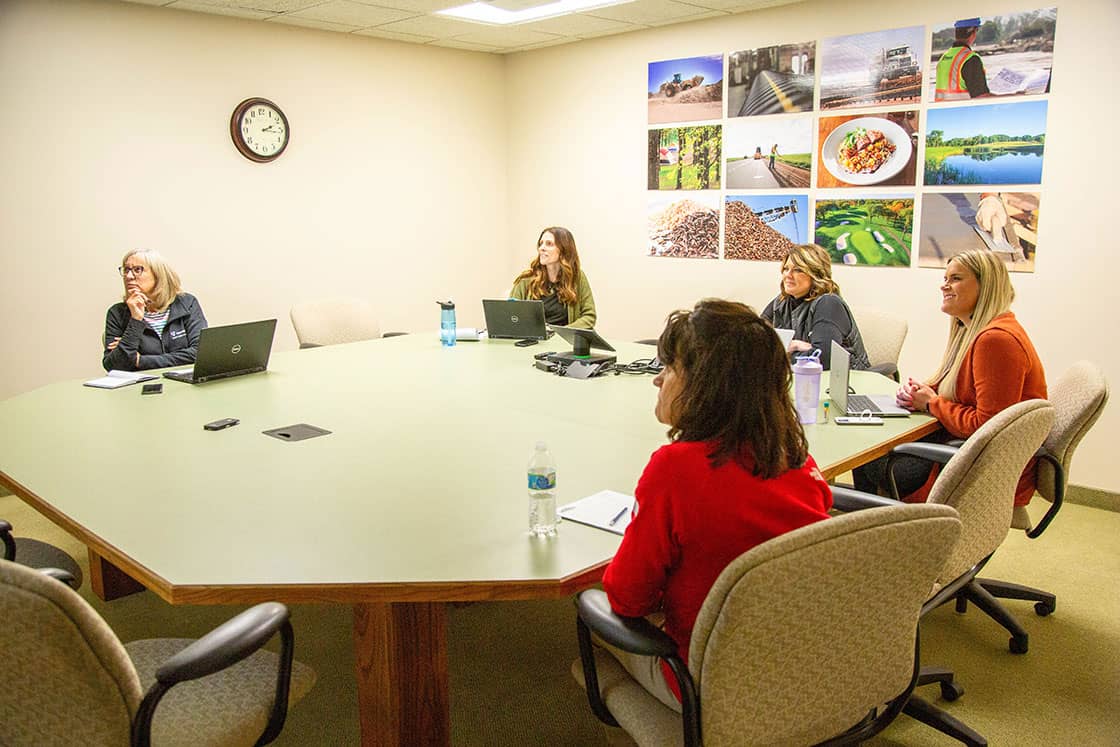 Five women at a conference room table looking at a PowerPoint.